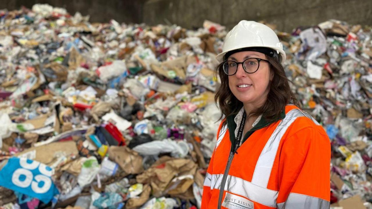 A woman wearing a white safety helmet and orange hi-vis jacket stands in front of a huge pile of waste in a warehouse. Items such as a large blue Co-op bag, plastic and paper can be seen.