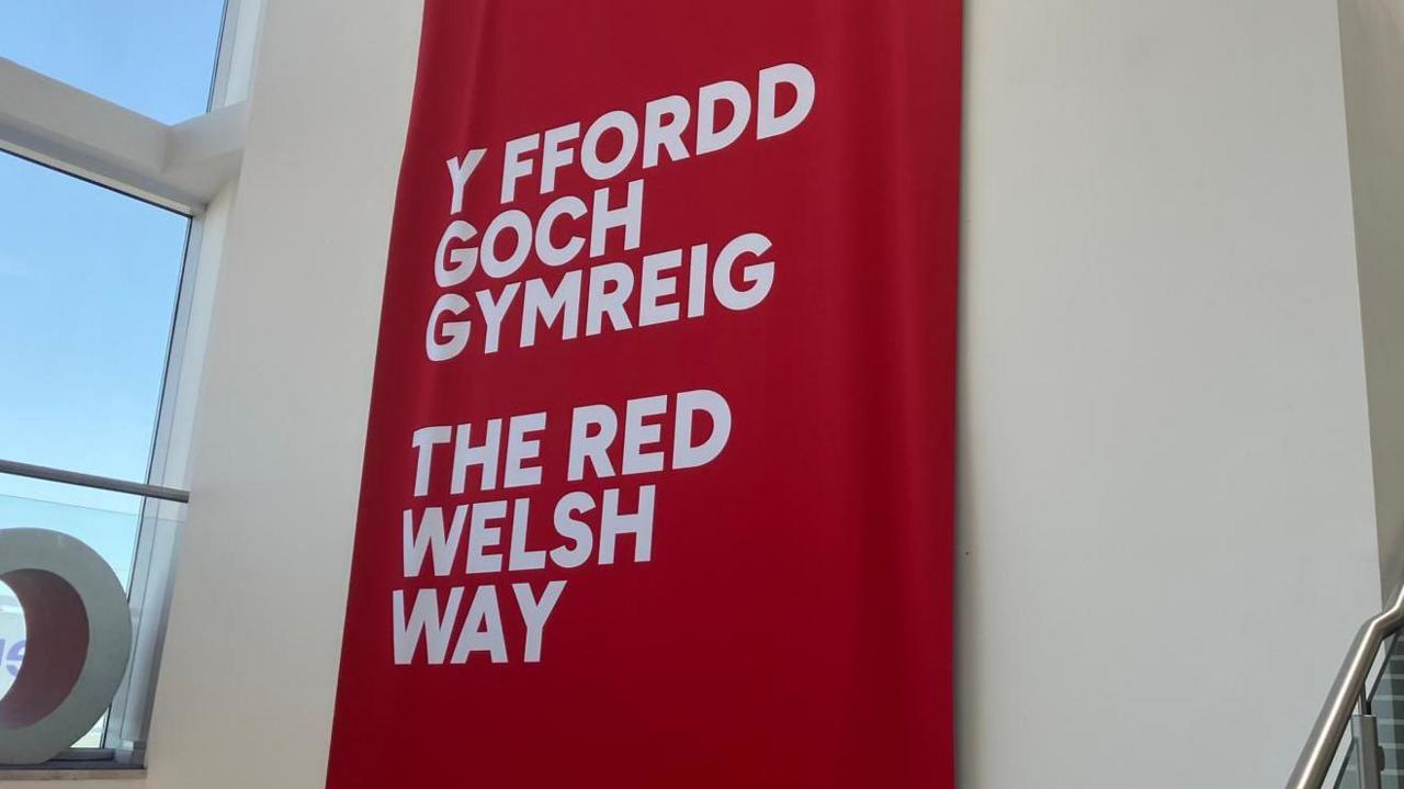 A red banner with "The Red Welsh Way" in English and Welsh at the front of Venue Cymru, with the blue skies over Llandudno beaming through a window to the left.