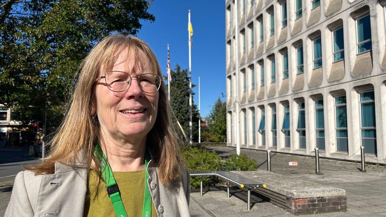 Labour councillor Ruth Williams wearing glasses and a green lanyard over a mustard sweater and a beige jacket. She is standing in front of Exeter's Civic Centre with flagpoles behind her.