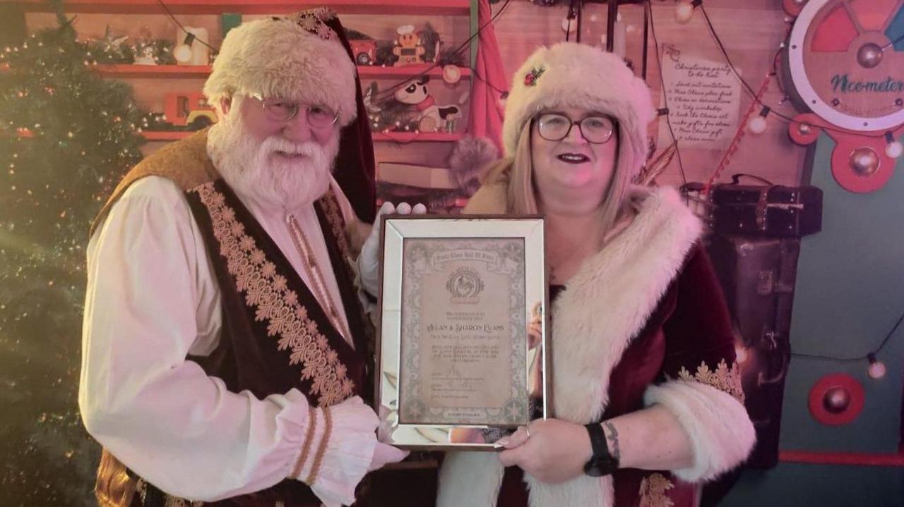 Allan and Sharon Evans, dressed in their Santa suits, jointly holding a certificate and standing in front of a Christmas tree and shelves of wooden toys