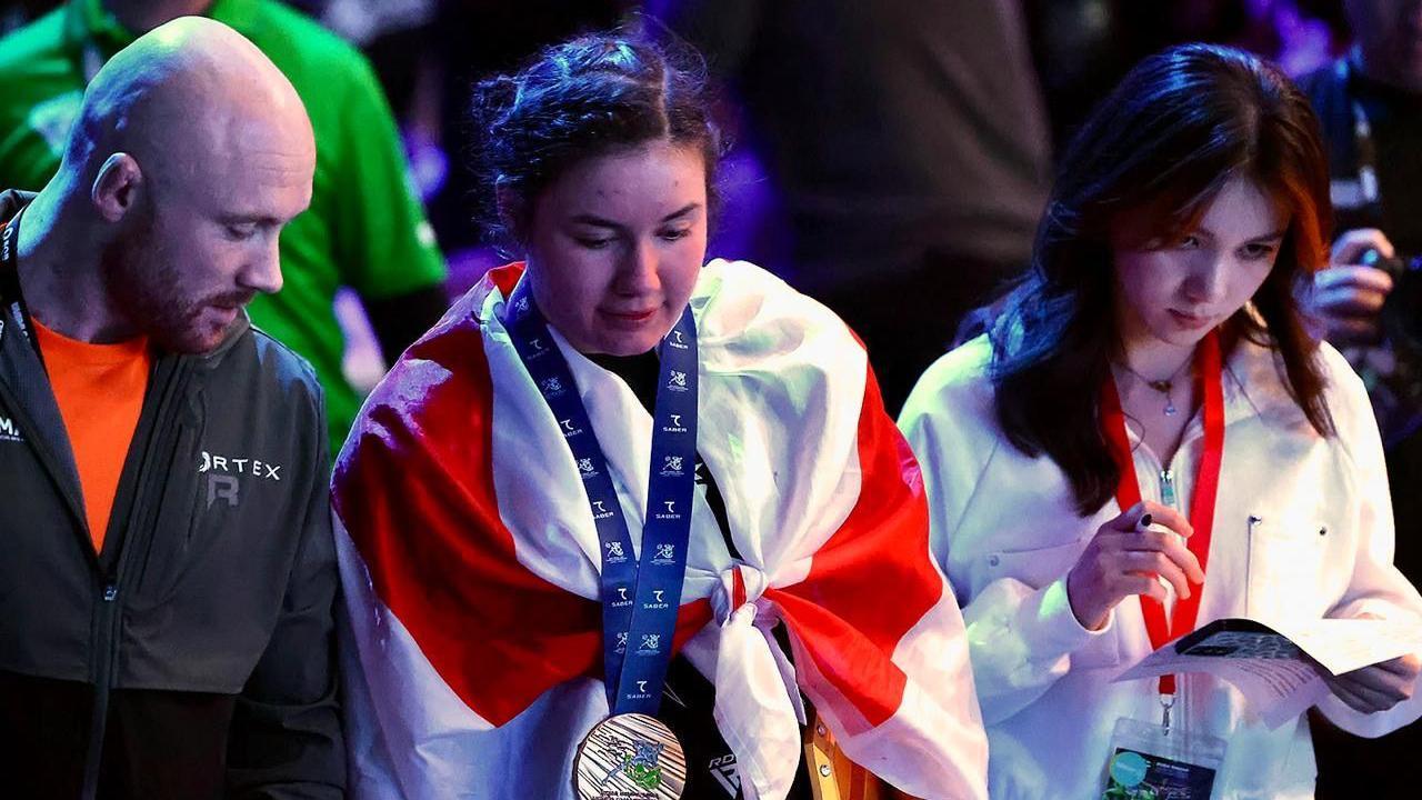 Alanna Pritchard walks next to a man and a woman. She wears a medal with a blue ribbon and a red and white flag over her shoulders.