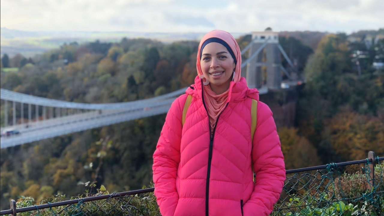 Dr Norah El-Gohary in a pink coat standing in front of Clifton Suspension Bridge.