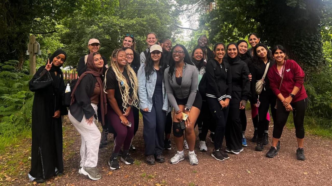 A group of women gather and pose for a photo on a path in the middle of a wooded area. They are of different backgrounds, some slightly crouch down for the photo or lean in, a couple make a positive V sign for the camera. Behind them are green bushes and trees with a brown open area under their feet.