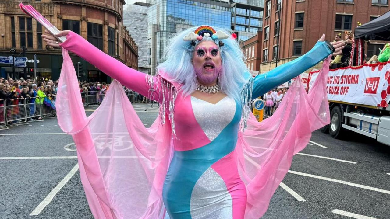 A person with a pink-dyed beard and wearing a pink, blue and white dress with a pink chiffon cape stands with their arms open wide in the Manchester Pride parade