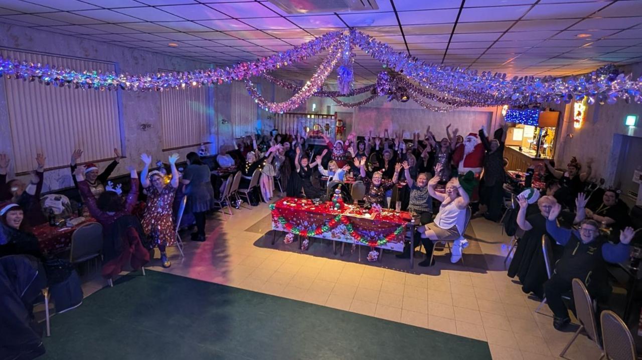 Dozens of women and a person dressed as Santa wave as they sit around tables in a function room decorated for Christmas.