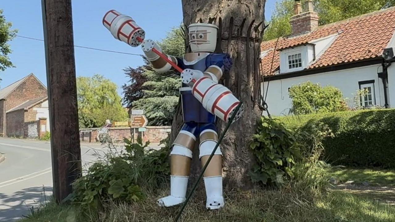 A character from Gladiators stands on a grass mound under a tree. The statue is holding a large stick used in one of the contests.