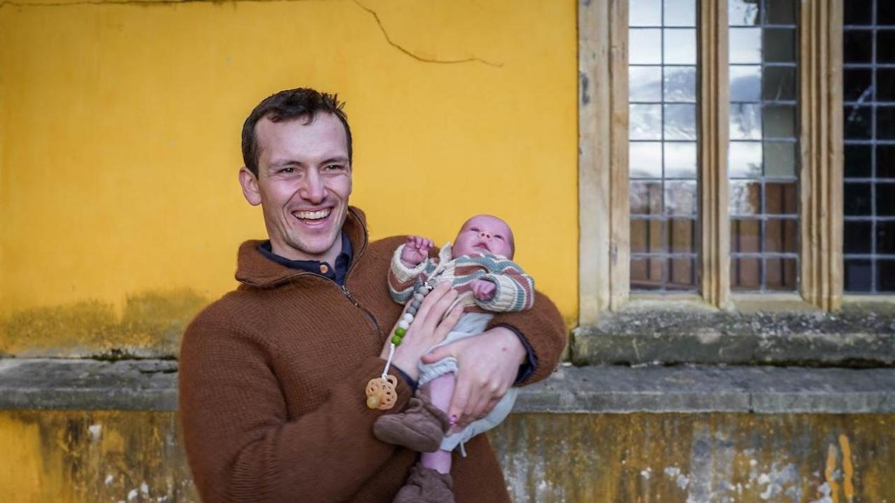 Richard is standing in front of a yellow building. He is holding a baby in his arms. He is wearing a brown jumper and has short cropped brown hair.