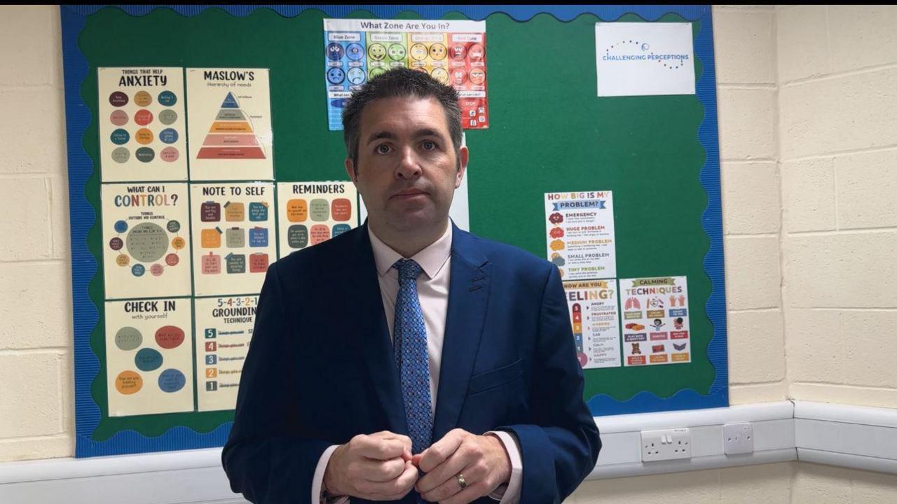 A man with dark-brown hair is wearing a blue suit jacket, white shirt, and blue tie. He is standing in front of a green wall-mounted information board with a blue border and posters on it.