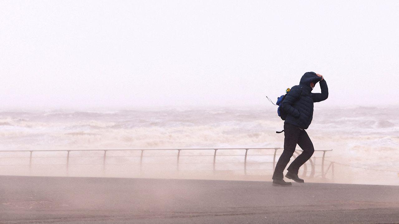A person walks along a promenade with a raging sea in the background, they're holding onto their hood as sea sprays up due to high winds, taken in Blackpool on 7 December.
