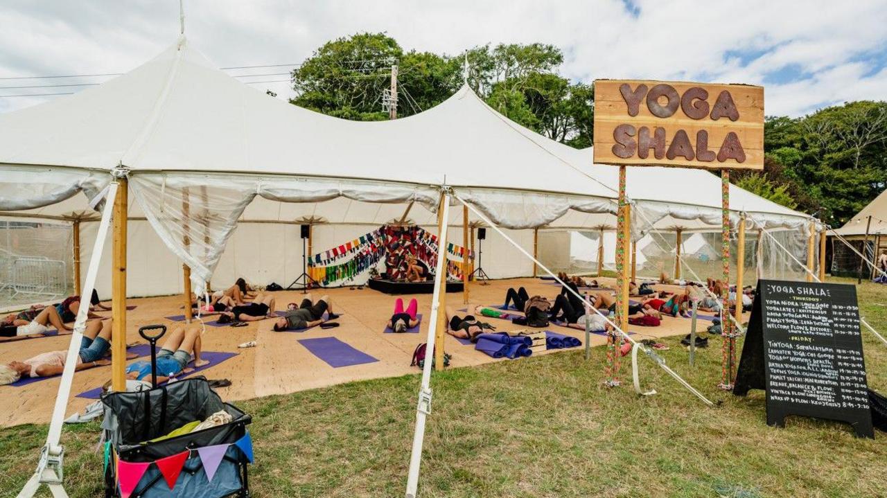 An open-sided marquee full of people laying on yoga mats. A wooden sign outside the tent reads - Yoga Shala.