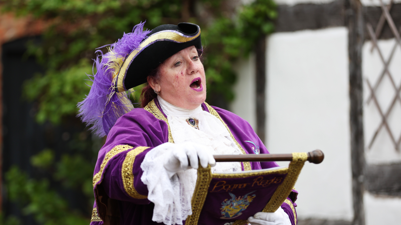 A woman in a purple town crier's outfit with her mouth open and holding a scroll in her hands