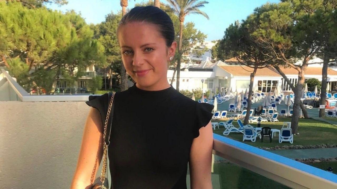 Hayley Morrison has dark hair which is tied back. She wears a black dress and carries a handbag on her shoulder. She is standing on a balcony at a holiday location with lots of deckchairs laid out on the grass behind her.