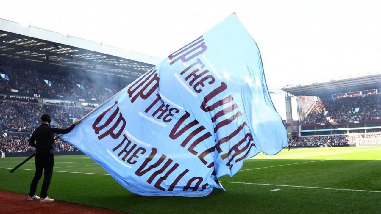 A blue Aston Villa flag is waved inside the stadium before a match. It says Up the Villa on it in maroon writing. Fans can be seen in grandstands around the pitch.