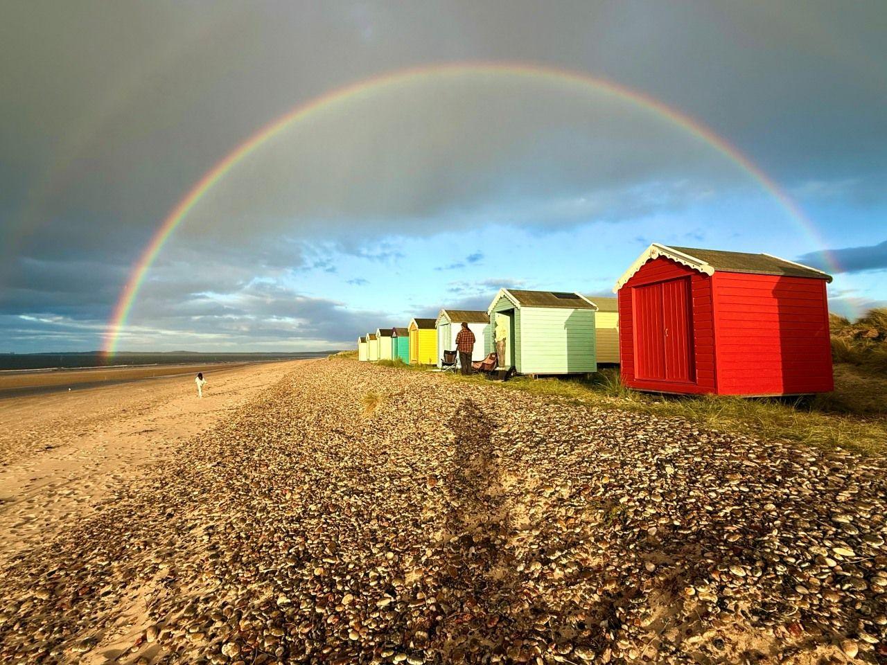 A row of bright colourful beach huts with a double rainbow overhead