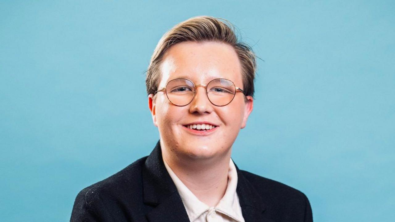 Oscar Brooker smiles, wearing glasses against a blue background