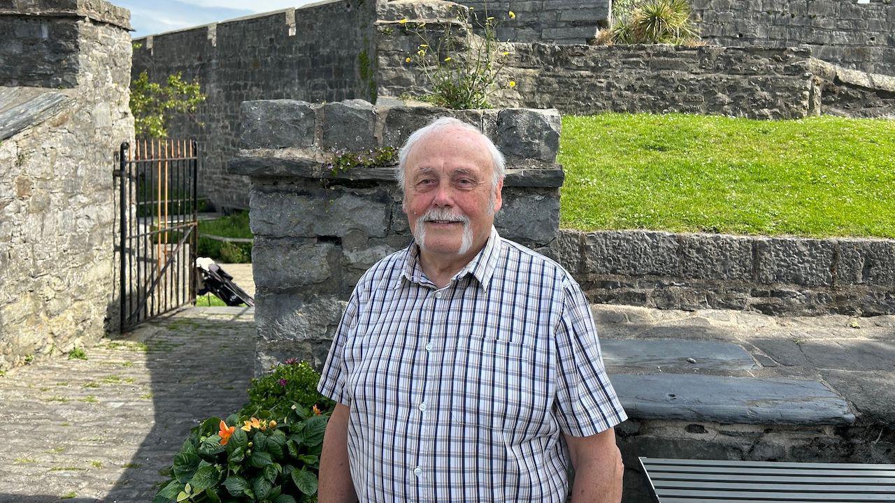 Tony Brown a man with white hair and a moustache stands smiling in front of the Castle in Castletown.