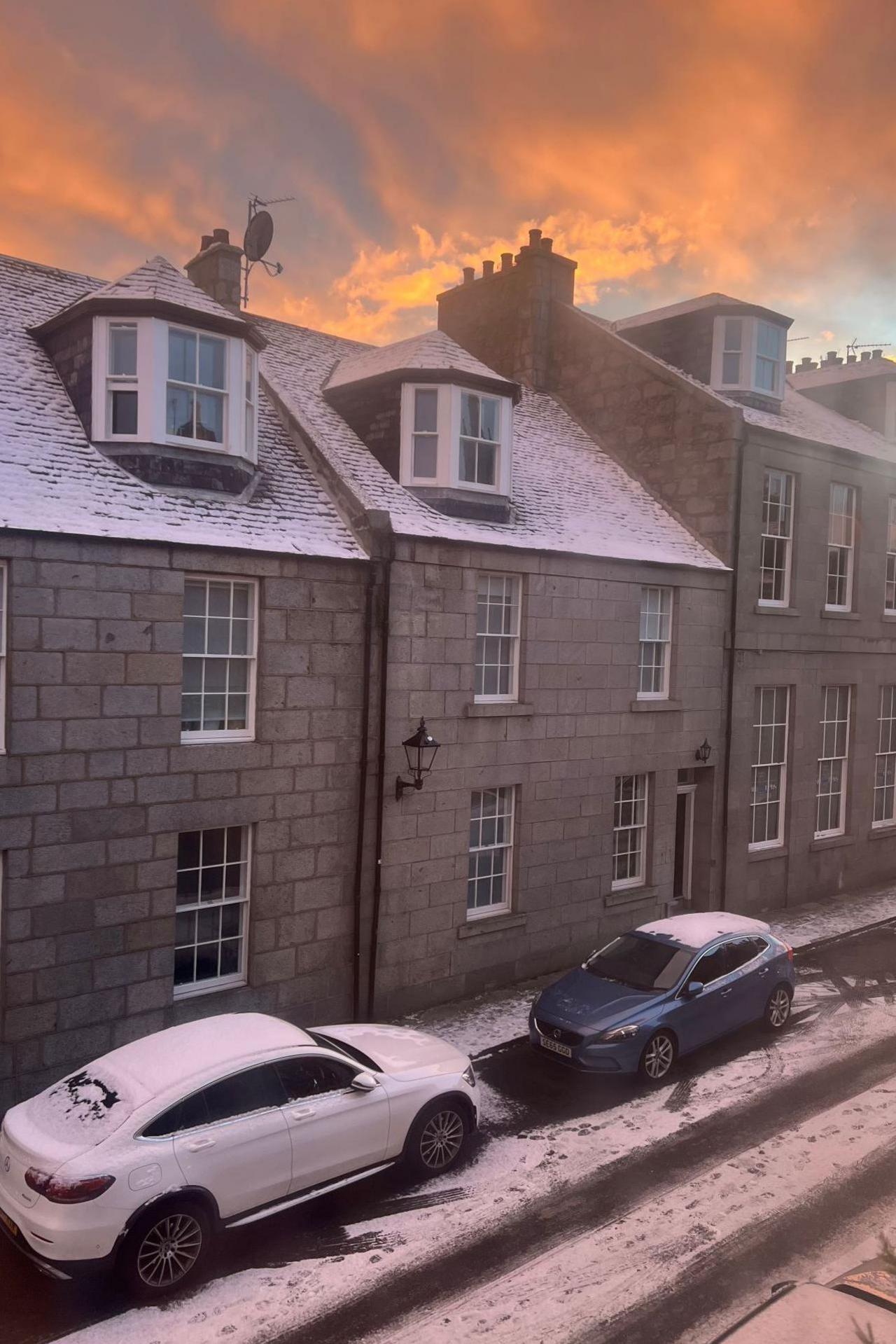 Snow lies on the road and on two cars parked in the street. There is a row of houses and the orange glow of sunrise behind them.