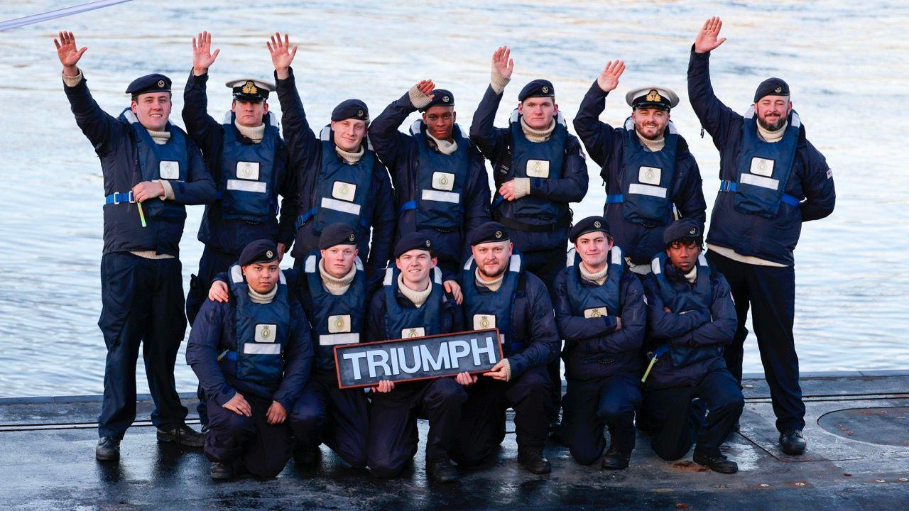 Royal Navy officers in two lines. The men in the front line are holding a sign which says "Triumph. They are stood on top of the submarine. In the background is the sea. It is a light blue colour.