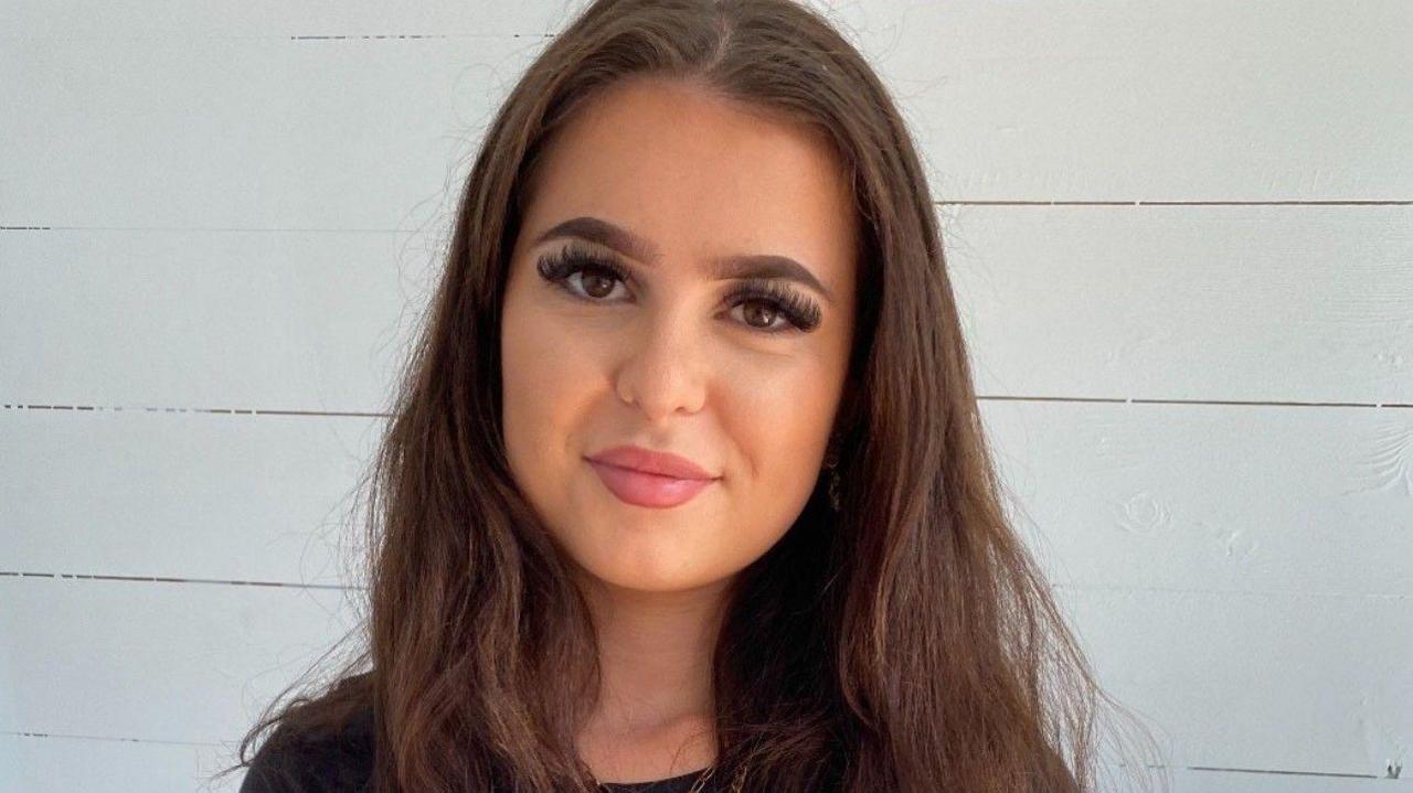 Labubu fan Victoria Calvert who has long brown hair and pink lipstick smiles at the camera next to a white wall