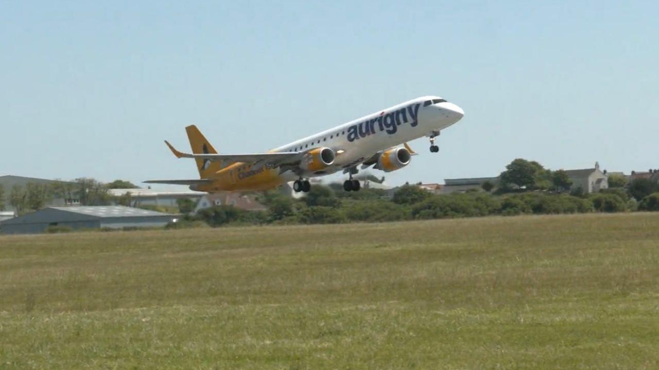 A yellow and white airplane with Aurigny logo on its side, setting off into the sky from the take off area at the airport. Blue skies and green grass visible.