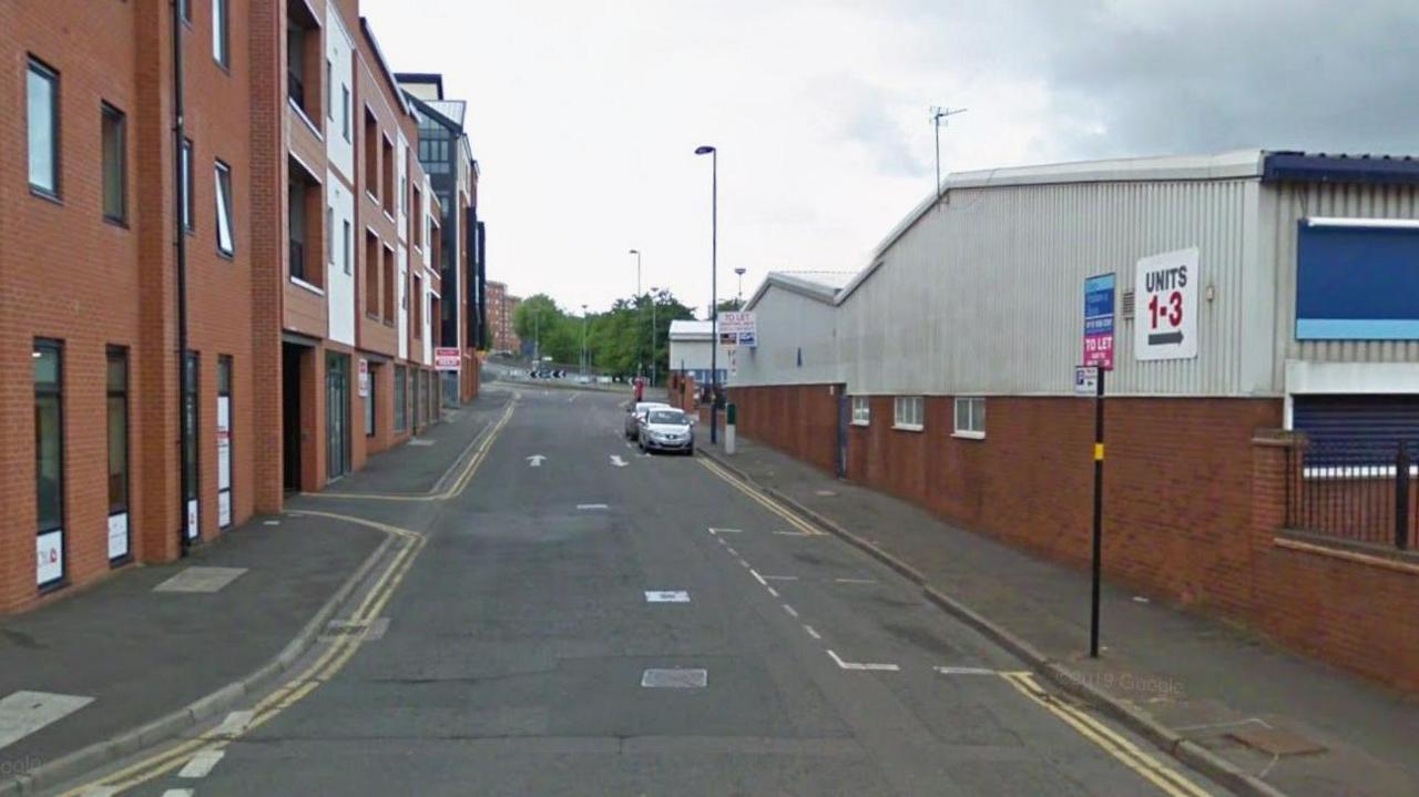 A street in Birmingham. It is quiet and there are only a couple of cars lining the road. On one side there are residential apartments. On the other there are industrial units. 