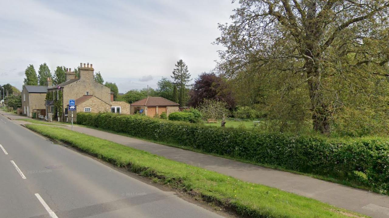 A road running alongside a grass verge, footpath and hedge into the village of Earith, with houses in the background.