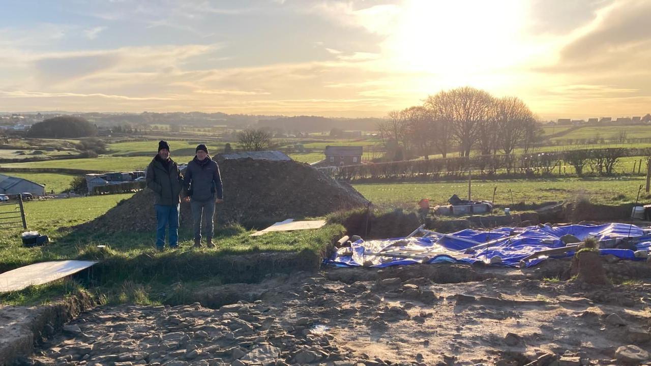 wider view of the rocky excavated site with mounds of rocks next to it. Bill and Chris stand near the edge of the excavated site. The sun rises behind trees on the farming area wit countryside behind.