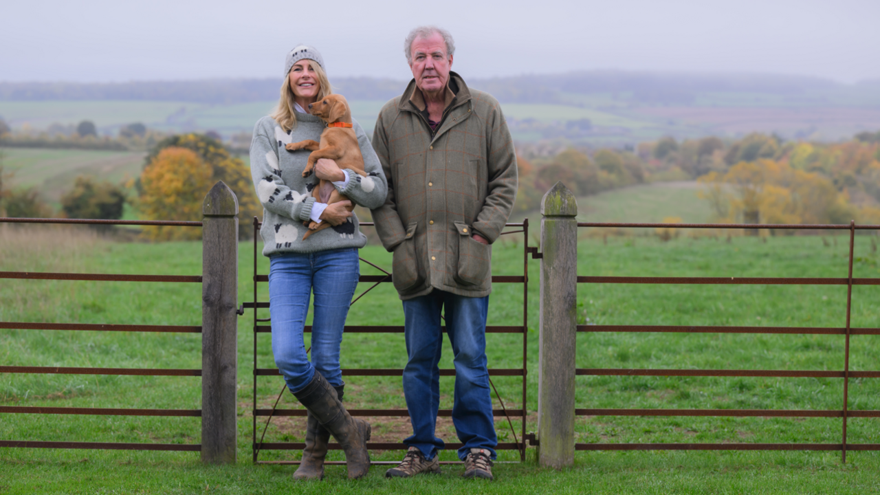 A woman with long blonde/brown hair and wearing a grey jumper is next to a man in a coat. They are both wearing blue jeans and standing by a gate in a field.