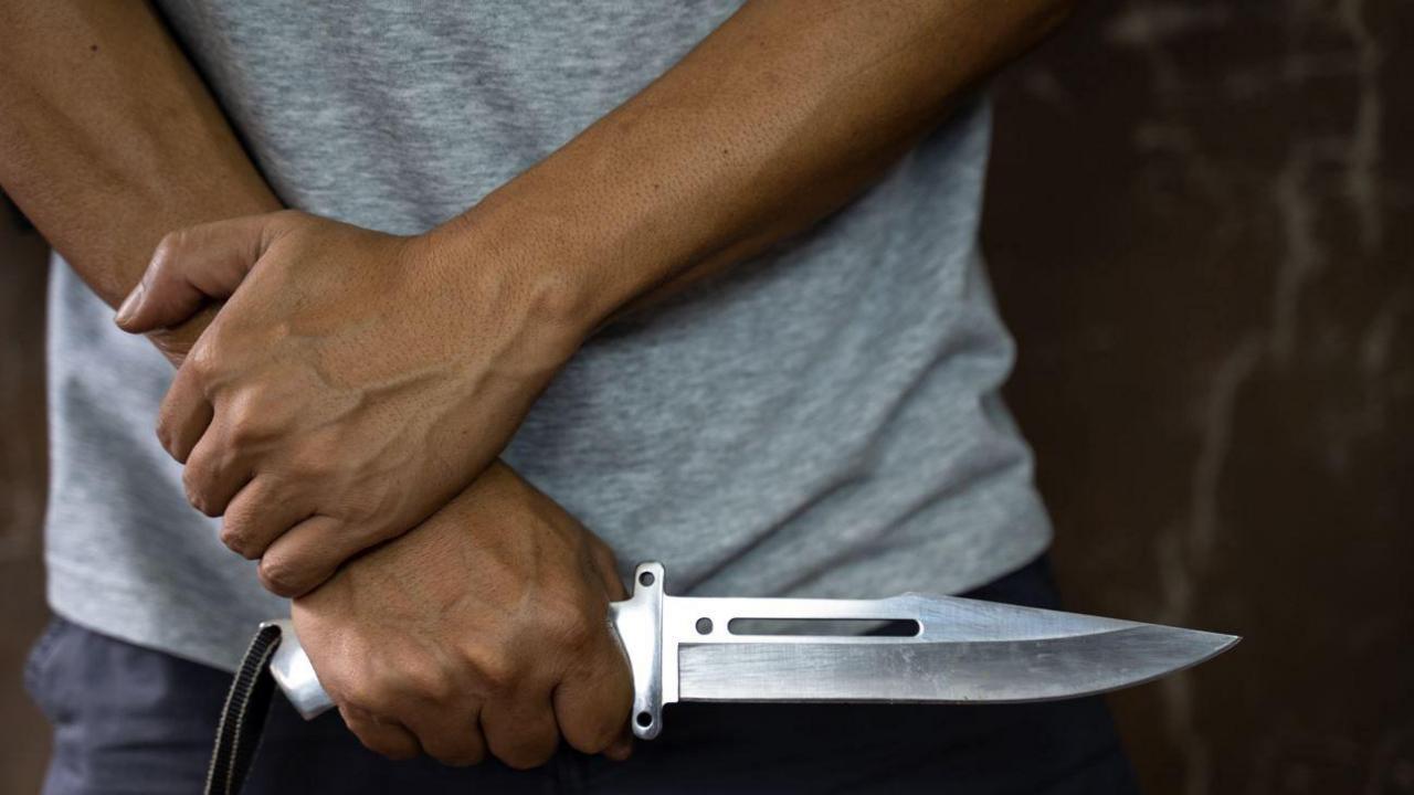 A silver bladed knife is being held by someone near their stomach with their other hand resting on their wrist. You can only see them waist down and they are wearing a grey top with black trousers.