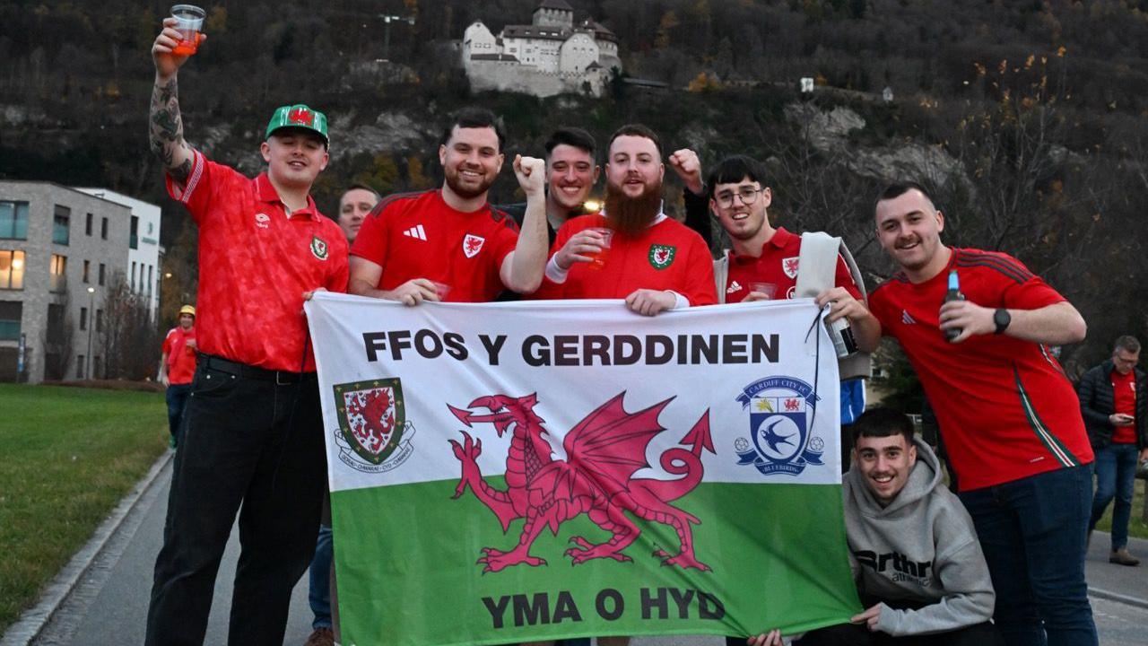 Grŵp o ddynion yn dal baner Cymru ac yn gwenu ar y camera. Maen nhw'n sefyll ar heol gydag adeilad gwyn ar ben bryn tu ôl iddyn nhw. Mae pump ohonyn nhw'n gwisgo crys-t coch tîm pêl-droed Cymru. Mae'r llall yn gwisgo siwmper lwyd.