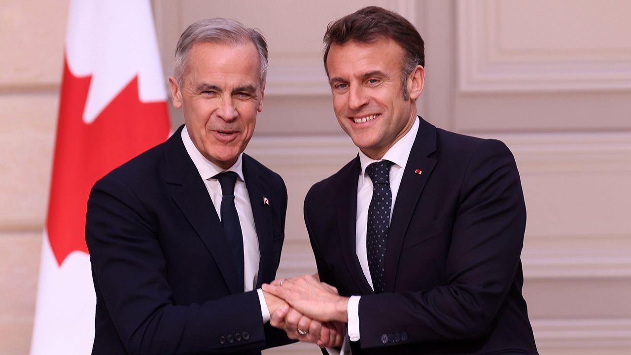 Mark Carney and Emmanuel Macron shake hands while smiling and looking at the camera in front of a Canadian flag during a press conference in Paris on Monday. They both wear a suit and tie.