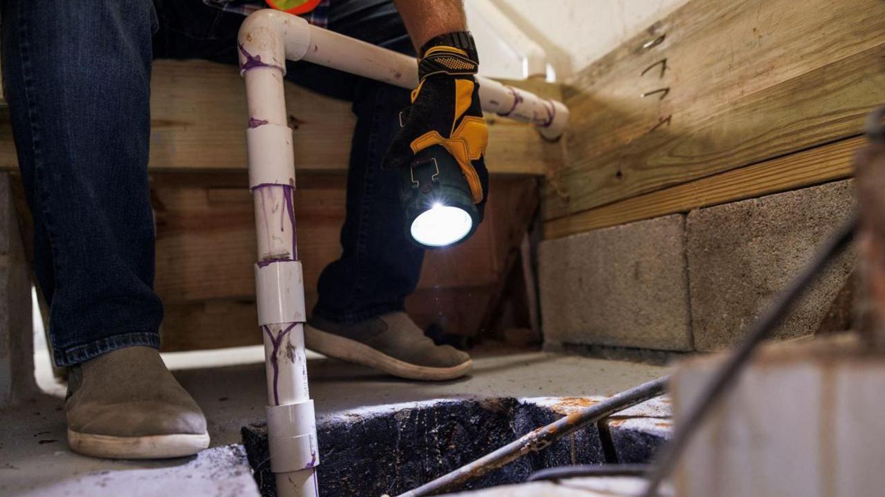 A square hole in concrete floor with a white pump coming out of it. A person wearing jeans and gloves is shining a torch down into the hole.