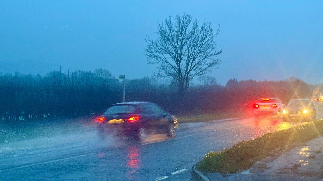 Two cars with lights on throw up spray as they drive down a road in the rain