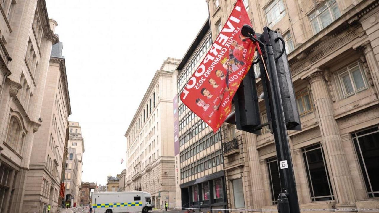 A red Liverpool FC flag hangs on a traffic light on the street. A police cordon is place and a police van is parked across the road. There are buildings on either side of the street. 