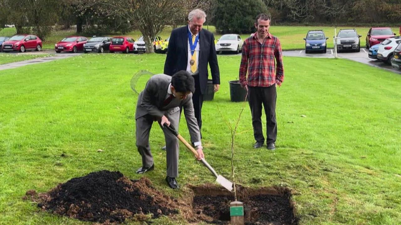 A man puts earth on top of a tree sapling. He is Japanese and is wearing a grey suit.
