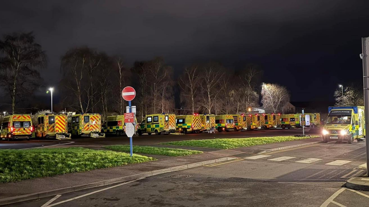 A row of ambulances lined up by a grass verge at night time. Another ambulance can be seen driving on a nearby road.