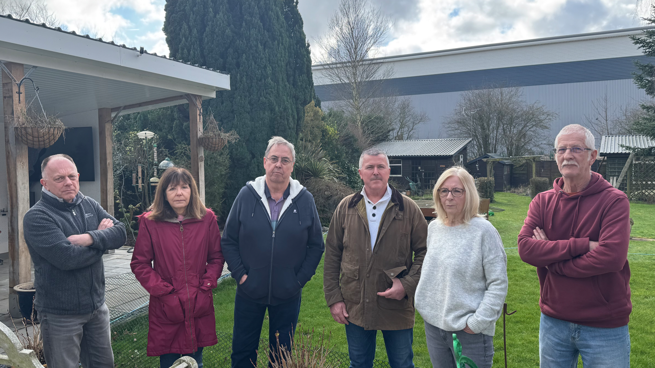 Four men and two women standing in a row in a back garden. The large warehouse can be seen in the background.