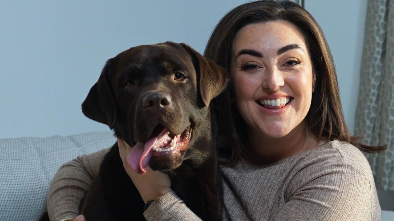 Bianca Perea with long brown hair smiling wearing a beige long sleeved t-shirt cuddling her brown Labrador dog as they sit on a cream sofa