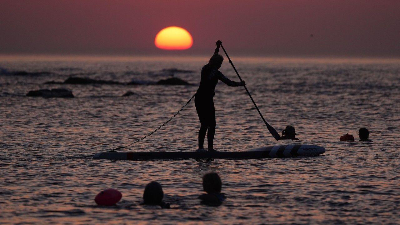 A person standing on a paddle board on the sea is silhouetted against a deep purple sky with the Sun rising on the horizon. Several swimmers are in the water too.