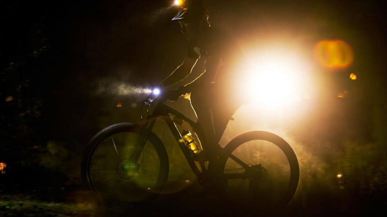 A silhouette of a man on a bicycle. It is a side view of him riding, with a bright light light in the background.