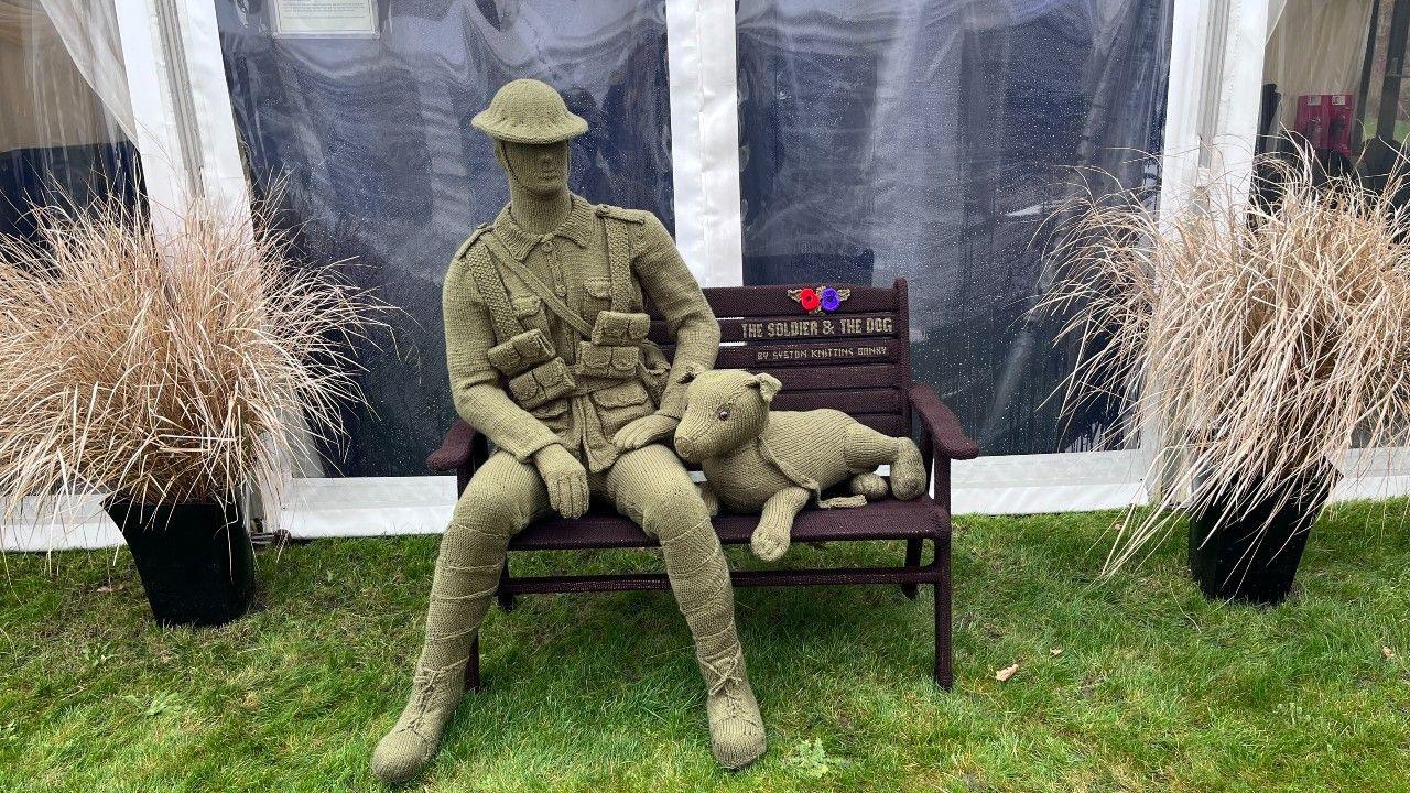 A khaki green knitted soldier figure, sitting on a bench in a relaxed pose, with a dog by his side, positioned within a garden area.