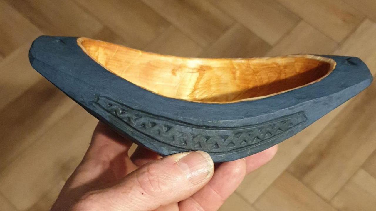 A hand holds a small, intricately carved wooden bowl with a dark exterior featuring geometric patterns and a smooth, light-coloured interior. The background shows wooden floor panels.