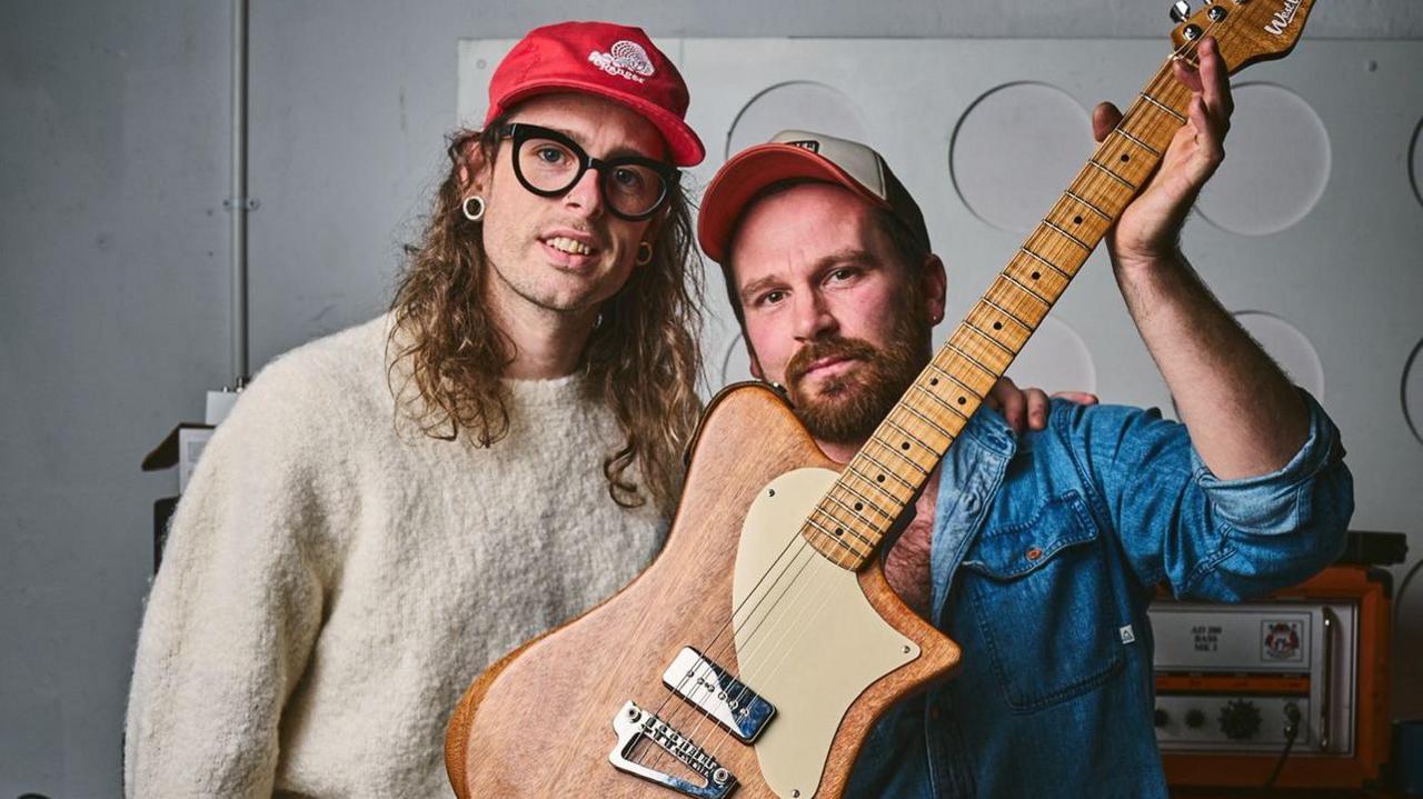 Idles' Lee Kiernan, on the left, is wearing a white jumper. On the left guitar maker Josh Stopford is wearing a blue denim shirt and holding a guitar