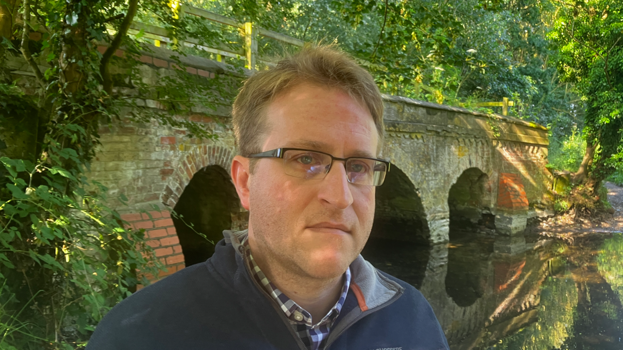 A man with short hair wearing glasses and a blue sweater standing in front of a bridge