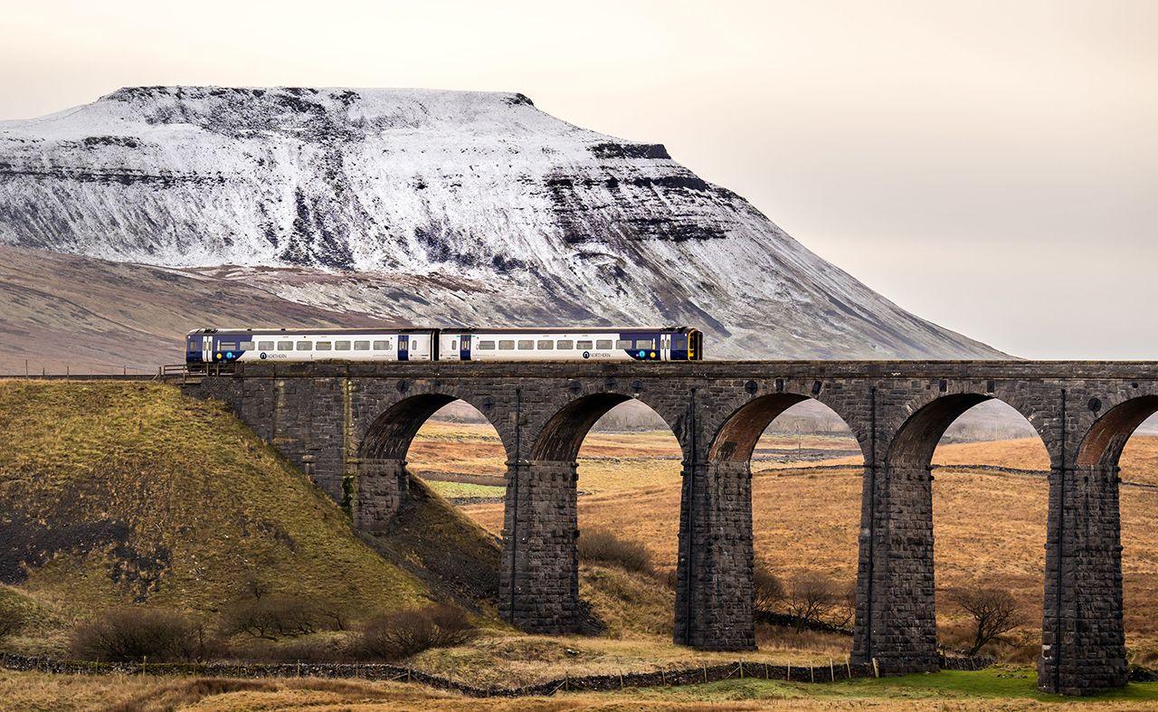 A Northern train travels over a stone bridge with snow on a rocky hill in the background in the Yorkshire Dales