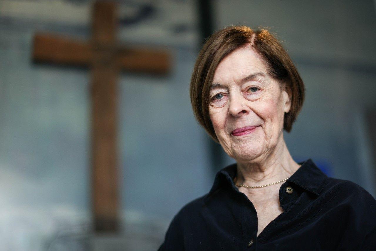 An 84-year-old woman wearing a black dress looks at the camera. Behind her is a large wooden cross mounted onto the wall.