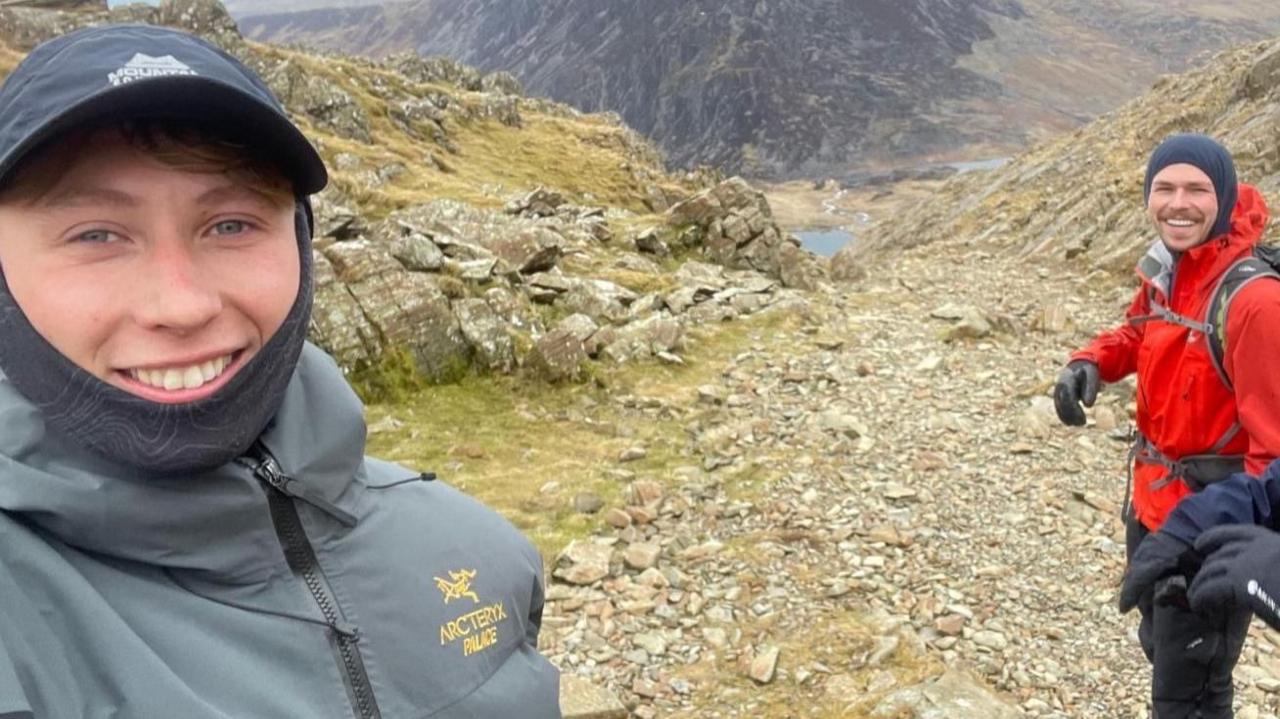 Mae dyn gyda het glas tywyll a siaced lwyd yn gwenu ar y camera. I'w chwith mae dyn mewn siaced goch, gyda sach gefn. Y tu ôl iddyn nhw mae mynydd llwyd creigiog a llyn glas yn y dyffryn islaw.