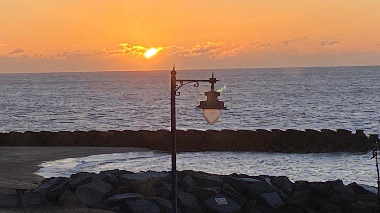 The sun sets in an orange sky over the sea with a decorative lamppost in the foreground