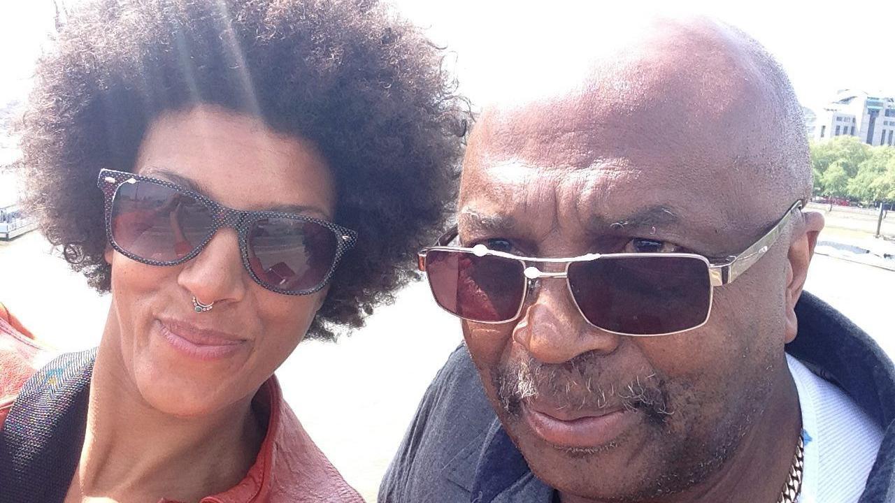 A dad and daughter stand together for a 'selfie' photograph, the woman has afro hair, a nose ring and wears sunglasses and the man has a greying moustache, bald head and wears sunglasses. 