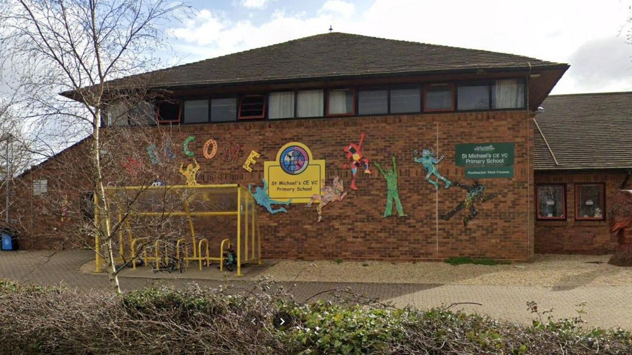 The outside of the school building near it's entrance with a few trees and bushes and a yellow bike locking shelter outside